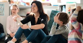 Eine Lehrerin sitzt mit Kindern in einem Kreis auf dem Boden und spricht mit ihnenen.